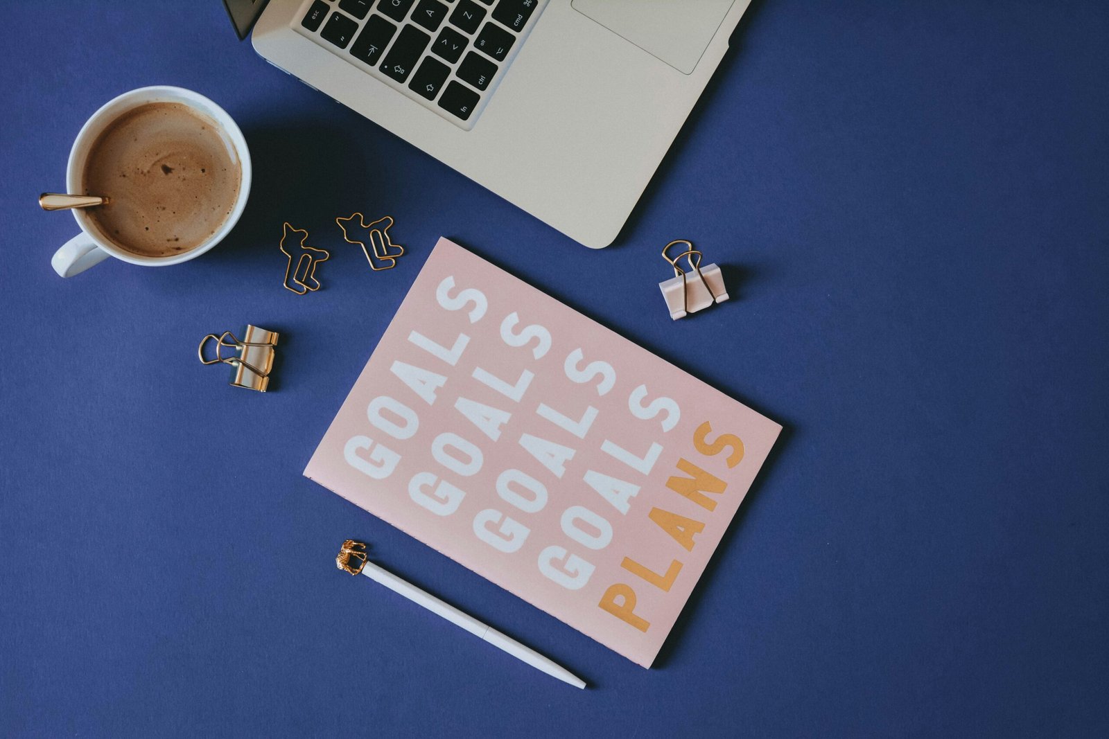Decorative photo of a blue table with a cup of coffee, a pink notebook with the word "Goals" and "Plans" on it, a white pencil, part of an apple laptop, and a few gold paperclips. 